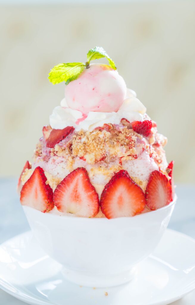 A bowl of creamy strawberry shortcake ice cream topped with fresh strawberries, crumbled shortcake pieces, and a dollop of whipped cream, served on a rustic wooden table.